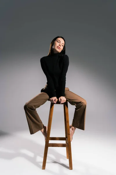 Sensual barefoot woman in brown pants and black sweater sitting on high stool on grey background — Stock Photo