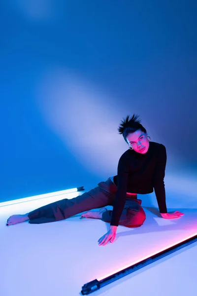 Longitud completa de mujer descalza con estilo posando cerca de lámparas fluorescentes sobre fondo azul - foto de stock