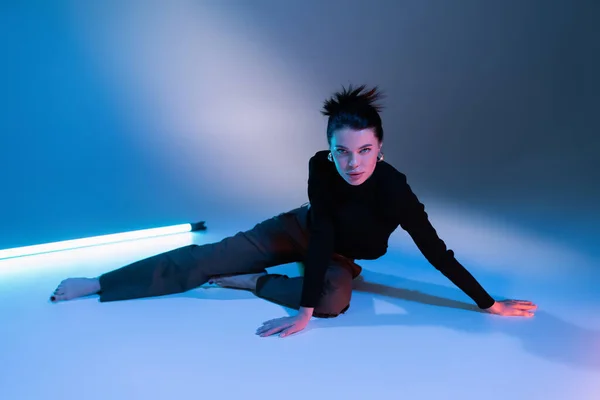 Brunette barefoot woman in black sweater and pants sitting near fluorescent lamp on blue background — Stock Photo