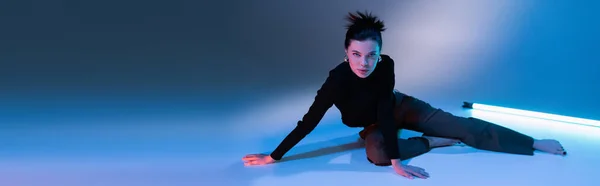 Pleine longueur de femme pieds nus en pull noir posant près de lampe fluorescente sur fond bleu, bannière — Photo de stock