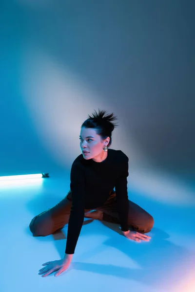 Donna alla moda in maglione nero seduto vicino alla lampada fluorescente e guardando lontano su sfondo blu — Foto stock