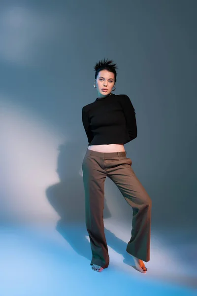 Full length of barefoot woman in black sweater and brown pants standing with hands behind back on grey background — Stock Photo