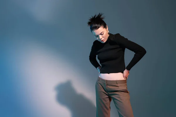 Young brunette woman in black sweater and brown trousers standing with hands on hips on grey background — Stock Photo