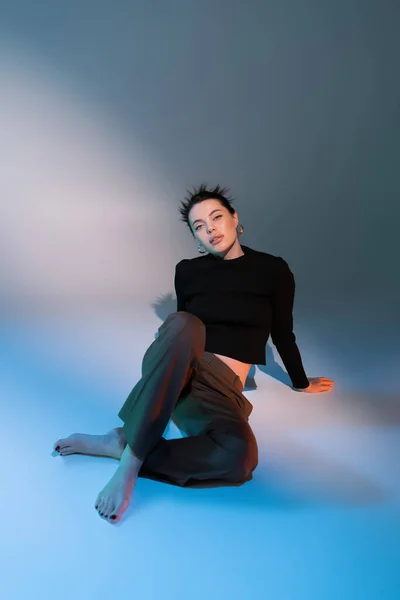 Longitud completa de sensual mujer descalza en pantalones marrones y suéter negro sentado sobre fondo gris y azul - foto de stock