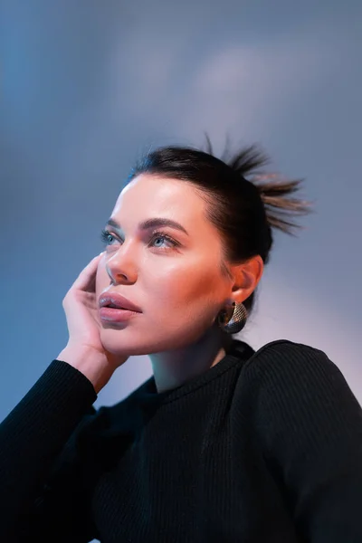 Retrato de mujer morena con maquillaje natural cogida de la mano cerca de la cara y mirando hacia otro lado sobre fondo azul - foto de stock