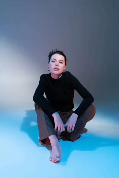 Comprimento total da mulher descalça elegante em camisola preta olhando para a câmera enquanto sentado em fundo cinza e azul — Fotografia de Stock