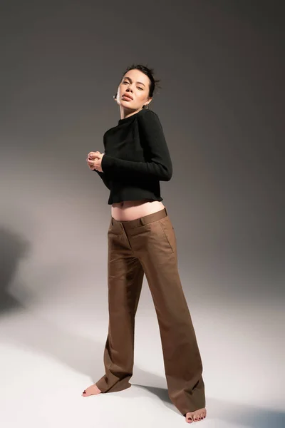 Brunette barefoot woman in brown trousers and black sweater standing on grey background — Stock Photo