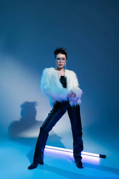 Full length of fashionable woman in white faux fur jacket posing near fluorescent lamp on blue background — Stock Photo