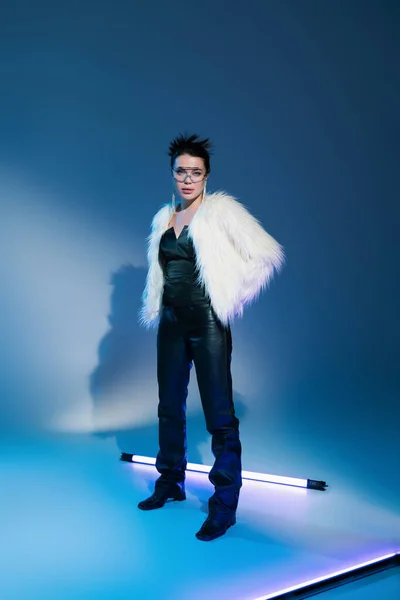 Full length of trendy woman in black leather pants and white furry jacket standing near fluorescent lamp on blue background — Stock Photo
