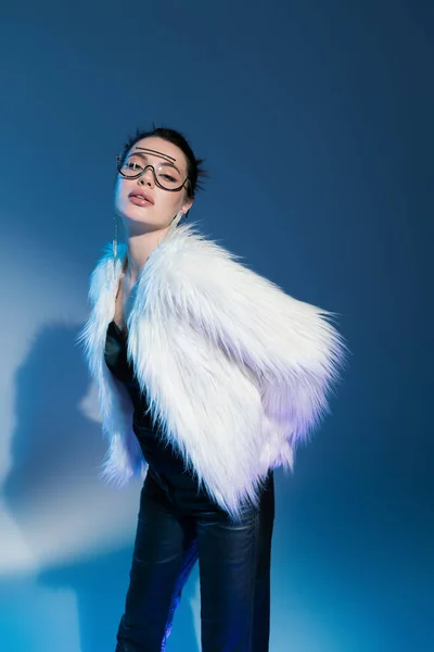 Young brunette woman in white faux fur jacket and transparent sunglasses looking at camera while posing on blue background — Stock Photo