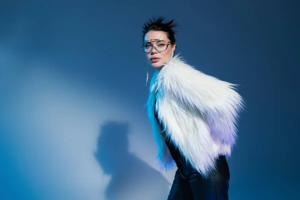 Young woman in transparent eyeglasses and fluffy faux fur jacket looking at camera on blue background — Stock Photo