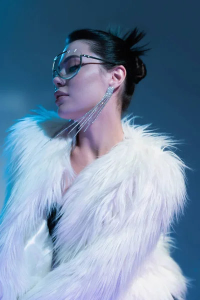 Brunette woman in transparent sunglasses and white faux fur jacket on blue background — Stock Photo