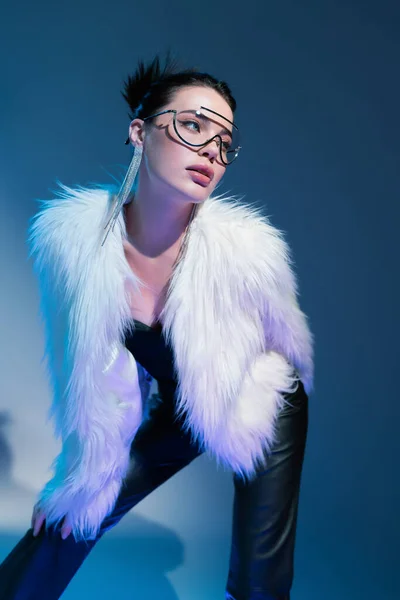 Modelo joven en gafas graduadas transparentes y chaqueta de piel sintética blanca mirando hacia otro lado sobre fondo azul - foto de stock