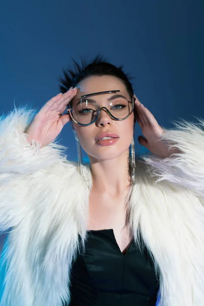 Fashionable woman in fluffy faux fur jacket and transparent sunglasses holding hands near face on blue background — Stock Photo