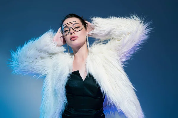 Pretty woman in black corset and white faux fur jacket posing with hands behind head on blue background — Stock Photo