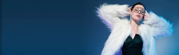 Brunette woman in transparent sunglasses and white furry jacket standing with hands behind head on blue background, banner — Stock Photo