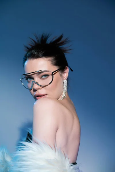 Portrait de femme sensuelle avec veste en fausse fourrure et épaule nue regardant la caméra en lunettes de soleil transparentes sur fond bleu — Photo de stock