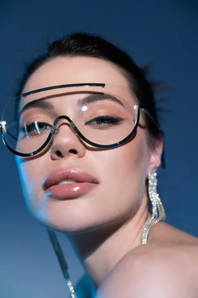 Nahaufnahme Porträt einer Frau mit Make-up und stylischer Sonnenbrille, die in die Kamera auf blauem Hintergrund blickt — Stockfoto