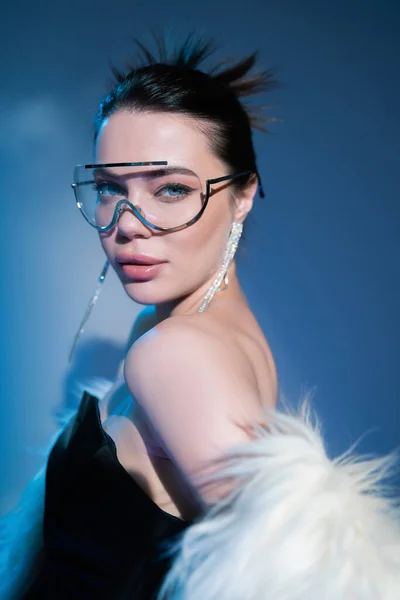 Mujer de moda en corsé negro y gafas de sol transparentes posando con chaqueta de piel sintética sobre fondo azul - foto de stock