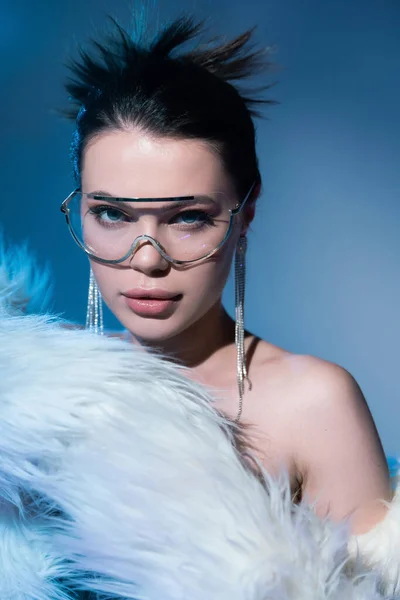 Porträt des Glamour-Models mit transparenter Sonnenbrille und glänzenden Ohrringen, die in der Nähe einer weißen Kunstpelzjacke auf blauem Hintergrund posieren — Stockfoto
