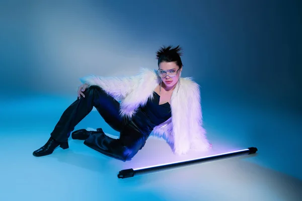 Trendy brunette woman in white faux fur jacket looking at camera near fluorescent lamp on blue background — Stock Photo