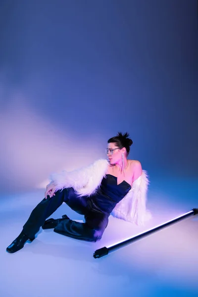 Glamour woman in black corset and white furry jacket sitting near fluorescent lamp on blue background — Stock Photo