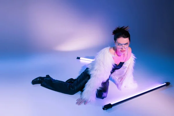 Femme à la mode en veste blanche en fausse fourrure posant près des lampes fluorescentes et regardant la caméra sur fond bleu et violet — Photo de stock