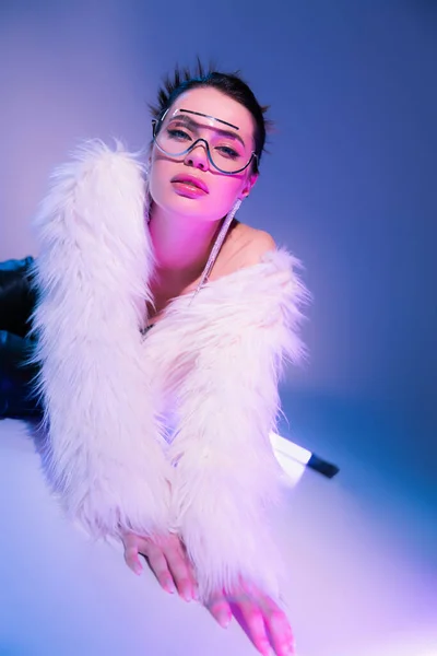 Trendy woman in white faux fur jacket lying near fluorescent lamp and looking at camera on blue and purple background — Stock Photo