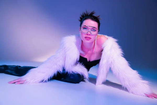 Séduisante femme en fausse fourrure blanche et lunettes de soleil transparentes posant sur fond bleu et violet — Photo de stock