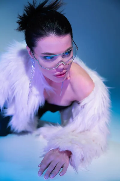 Mujer elegante en gafas de sol de moda y chaqueta de piel sintética posando sobre fondo azul con luz púrpura - foto de stock