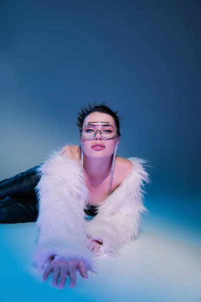 Glamour woman in white faux fur jacket and transparent eyeglasses lying on blue background — Stock Photo