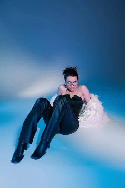 Longitud completa de la mujer con estilo en chaqueta de piel sintética blanca y pantalones de cuero negro posando sobre fondo azul - foto de stock