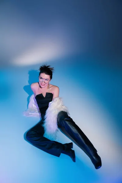 Mujer sonriente en pantalones de cuero negro y chaqueta de piel sintética de moda posando sobre fondo azul - foto de stock