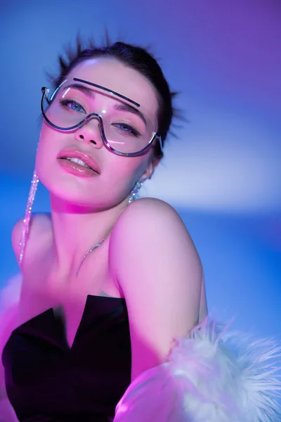 Jeune femme sensuelle en lunettes de soleil à la mode et boucles d'oreilles brillantes regardant la caméra sur fond bleu et violet — Photo de stock
