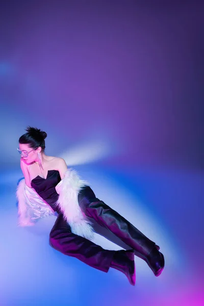 Longitud completa de la mujer morena en pantalones de cuero negro y chaqueta de piel sintética blanca acostado sobre fondo púrpura - foto de stock