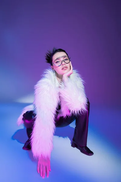 Модна жінка в прозорих окулярах і біла пухнаста куртка дивиться на камеру на фіолетовому фоні — стокове фото