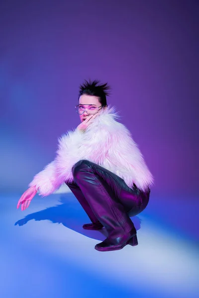 Full length of brunette model in black leather pants and white furry jacket looking at camera on purple background — Stock Photo