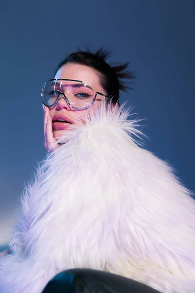 Sinnliches brünettes Modell in weißer Kunstpelzjacke und transparenter Brille, die das Gesicht auf blauem Hintergrund berührt — Stockfoto