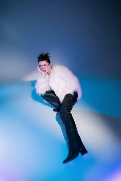 High angle view of fashionable woman in white furry jacket looking at camera while posing on blue background — Stock Photo