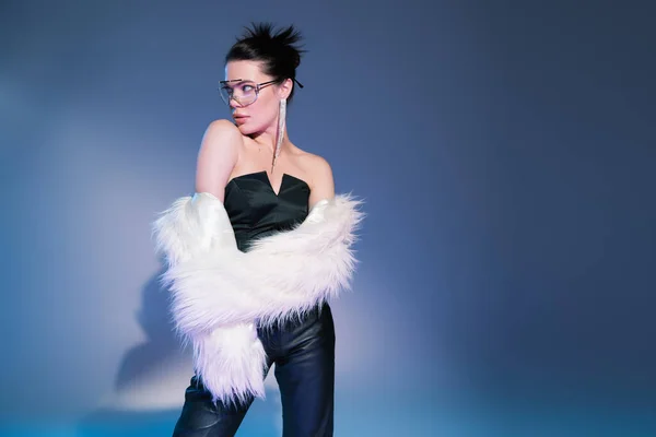 Glamour model in black corset with leather pants looking away while posing in white faux fur jacket on blue background — Stock Photo