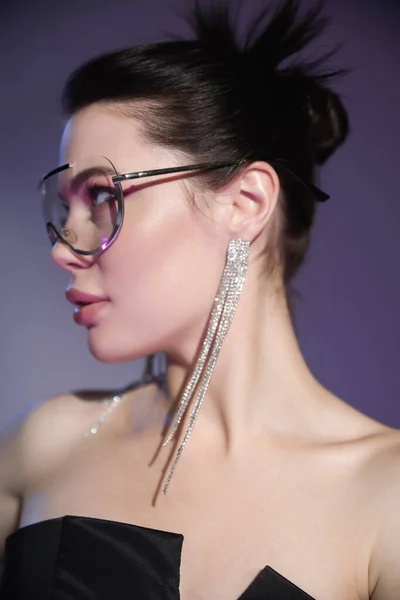 Jeune femme brune en boucles d'oreilles brillantes et lunettes de soleil à la mode regardant loin sur fond violet — Photo de stock