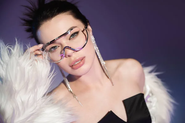 Jeune femme en fausse fourrure blanche touchant des lunettes transparentes sur fond violet — Photo de stock