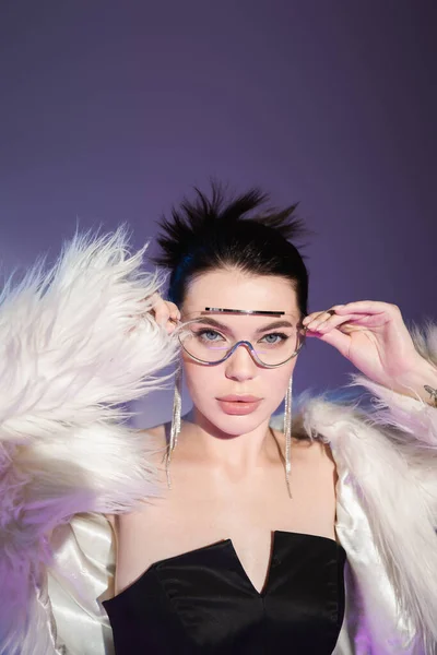 Glamour model in white faux fur jacket adjusting transparent sunglasses on purple background — Stock Photo