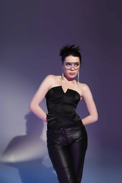 Brunette woman in fashionable eyeglasses and black corset posing with hands behind back and looking away on purple background — Stock Photo