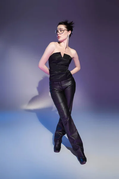 Longueur totale de glamour femme en pantalon en cuir et corset noir posant avec les mains derrière le dos sur fond violet — Photo de stock