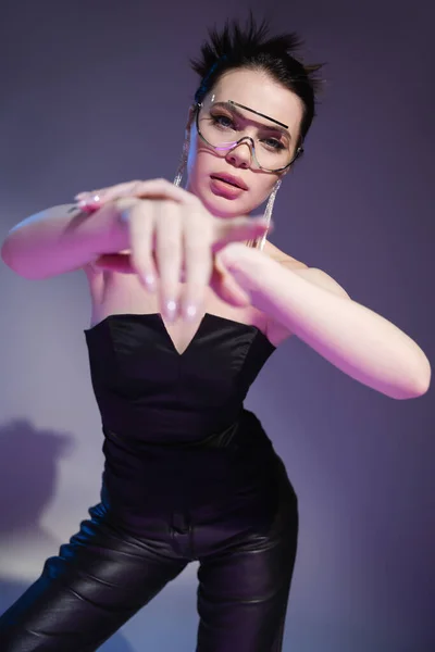 Brunette woman in black corset and trendy sunglasses looking at camera and pointing with finger on purple background — Stock Photo