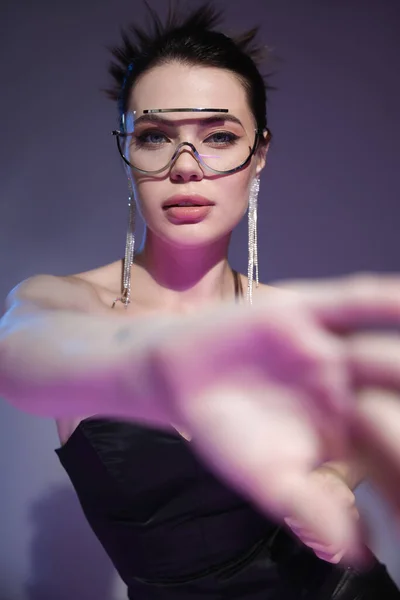 Glamour-Frau mit durchsichtiger Sonnenbrille blickt in die Kamera in der Nähe verschwommener ausgestreckter Hand auf lila Hintergrund — Stockfoto