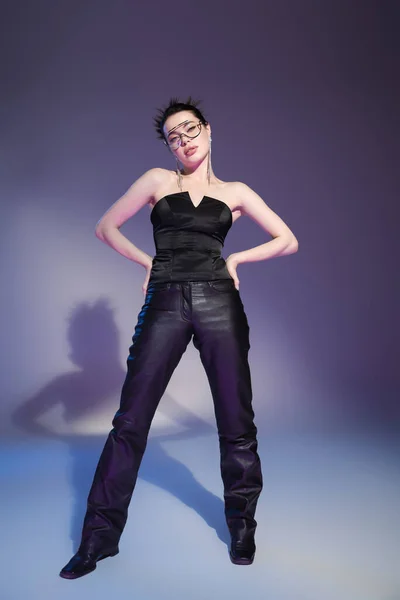 Piena lunghezza della donna alla moda in pantaloni di pelle nera e corsetto posa con le mani sui fianchi su sfondo viola — Foto stock
