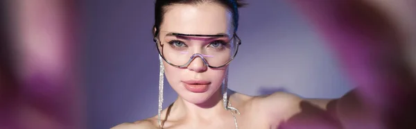 Portrait de jeune femme en lunettes de soleil transparentes et boucles d'oreilles brillantes regardant la caméra sur fond violet, bannière — Photo de stock