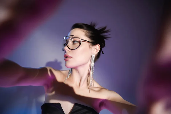 Brunette woman in trendy sunglasses and shiny earrings looking away on blurred purple background — Stock Photo